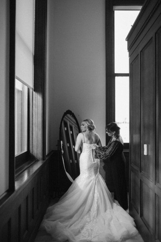 A bride in a long, elegant gown stands near a window as another woman helps her adjust the dress. A large mirror and wooden paneling are visible in the softly lit, black-and-white scene. by Justin Salem Meyer