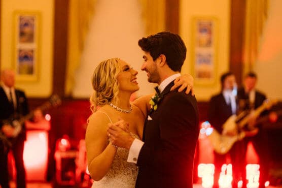 A bride and groom share a joyful first dance at their wedding reception, smiling at each other. A live band plays in the background under warm, golden lighting. by Justin Salem Meyer