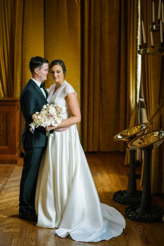 A couple stands close together indoors; one wears a black suit and bow tie, the other a white wedding dress holding a bouquet. They stand on wooden floors with gold curtains and candelabras in the background. by Justin Salem Meyer