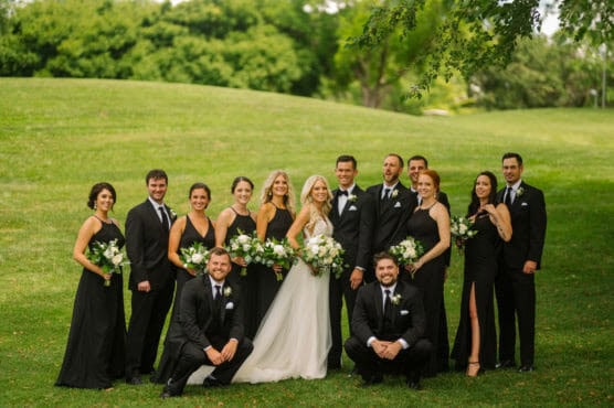 A wedding party of twelve people poses outdoors on grass. The bride stands in the center in a white gown, surrounded by bridesmaids in black dresses and groomsmen in black suits, all holding bouquets and smiling. Trees and greenery are in the background. by Justin Salem Meyer