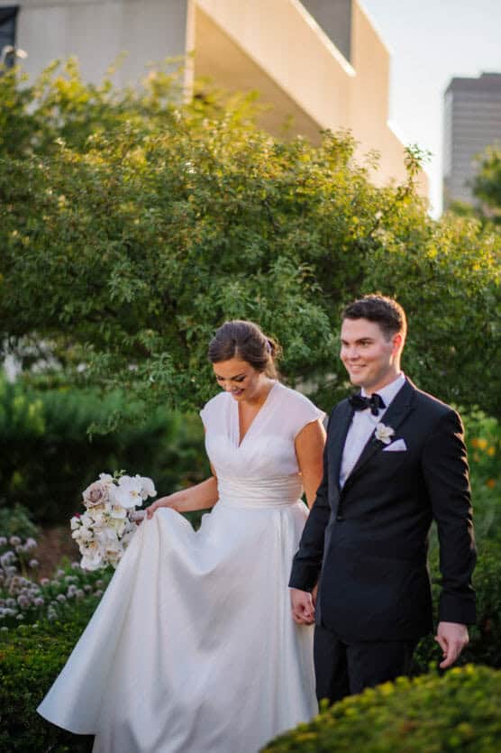 A bride in a white gown holds a bouquet of white and pale flowers while walking hand in hand with a groom in a black tuxedo through a garden, both smiling softly. by Justin Salem Meyer