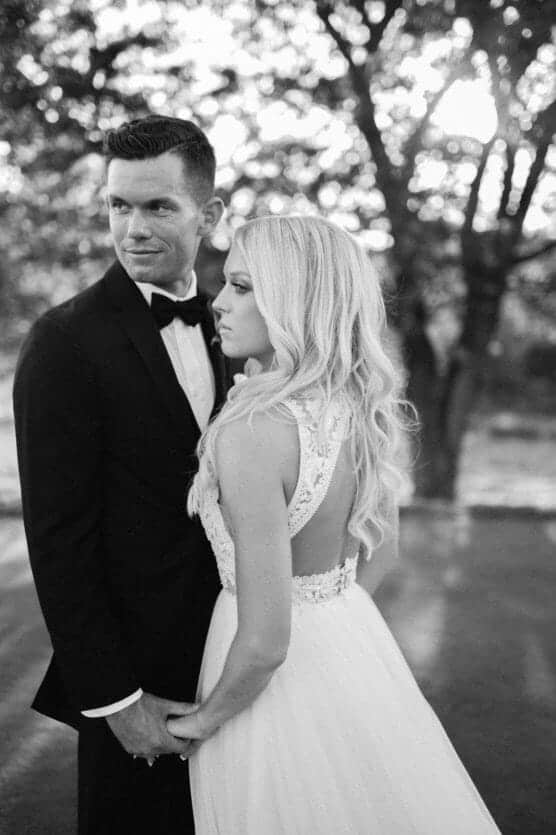 A bride in a sleeveless lace wedding dress and a groom in a black tuxedo stand together outdoors, holding hands and looking in different directions, with sunlight filtering through trees in the background. by Justin Salem Meyer