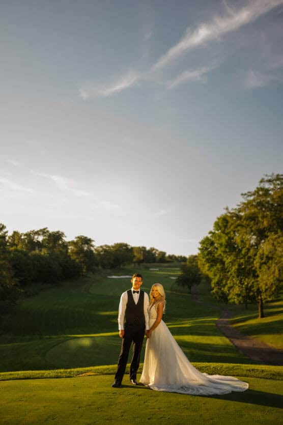 A bride and groom stand together on a green golf course at sunset, holding hands and smiling. The bride wears a long white dress and the groom is in a tuxedo. Trees and a clear sky are in the background. by Justin Salem Meyer