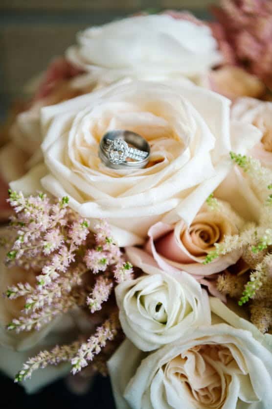 A close-up of a bridal bouquet featuring cream and blush roses with two silver wedding rings nestled in the center of a large white rose. by Justin Salem Meyer
