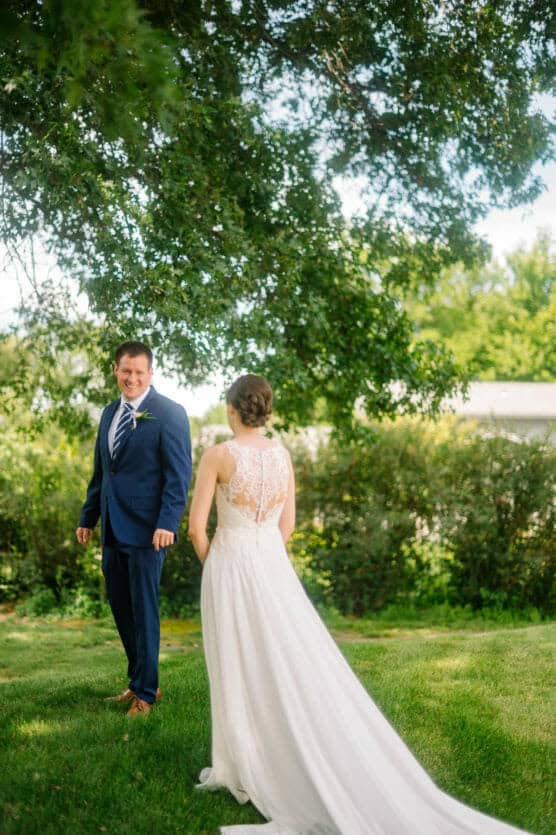 A bride in a white lace dress walks toward a smiling groom in a blue suit, standing on green grass under a large leafy tree on a sunny day. by Justin Salem Meyer