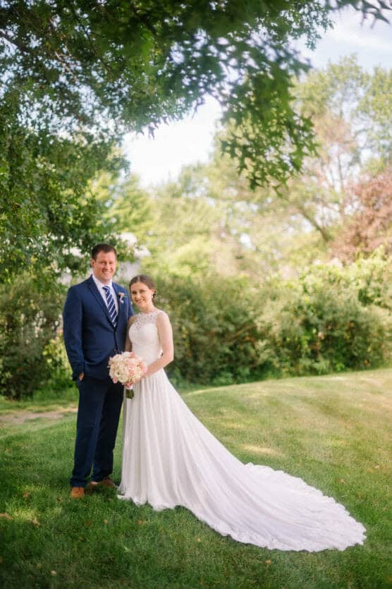 A bride in a white gown holding a bouquet stands beside a groom in a dark suit on a grassy lawn, surrounded by green trees and bushes on a sunny day. by Justin Salem Meyer