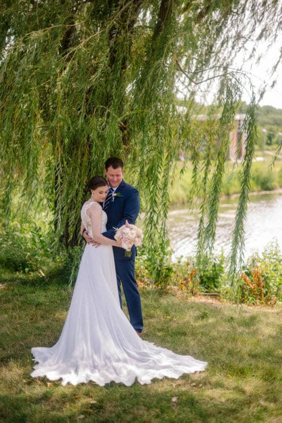A bride in a flowing white gown and a groom in a dark blue suit embrace under a willow tree by a pond, with sunlight filtering through the leaves. The bride holds a bouquet of flowers and both look peaceful and happy. by Justin Salem Meyer