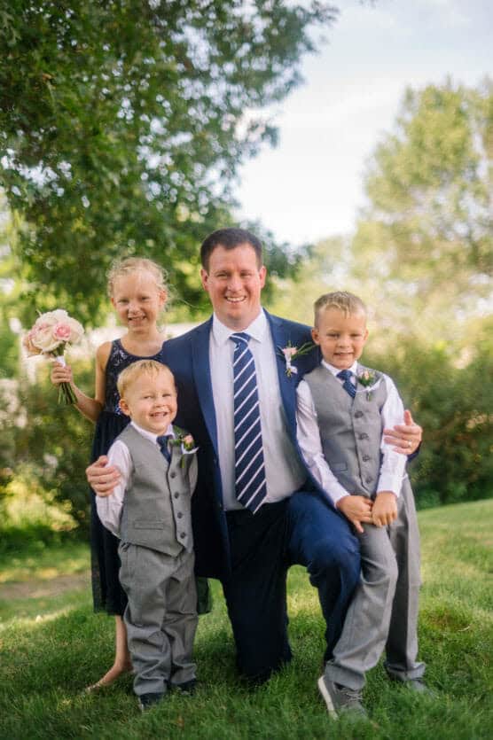 A man in a suit and striped tie kneels on grass outdoors, smiling with three young children dressed formally. A girl holds a bouquet of pink roses. Green trees and sunlight fill the background. by Justin Salem Meyer