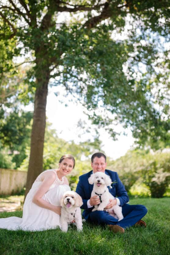 A smiling bride and groom sit on grass under a large tree, each holding a small white dog. The bride wears a white dress, and the groom wears a blue suit. Sunlight filters through the leaves in a lush, green outdoor setting. by Justin Salem Meyer