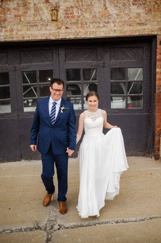 A smiling bride in a white dress and a groom in a blue suit walk hand-in-hand on a sidewalk in front of a rustic brick building with dark garage doors. by Justin Salem Meyer