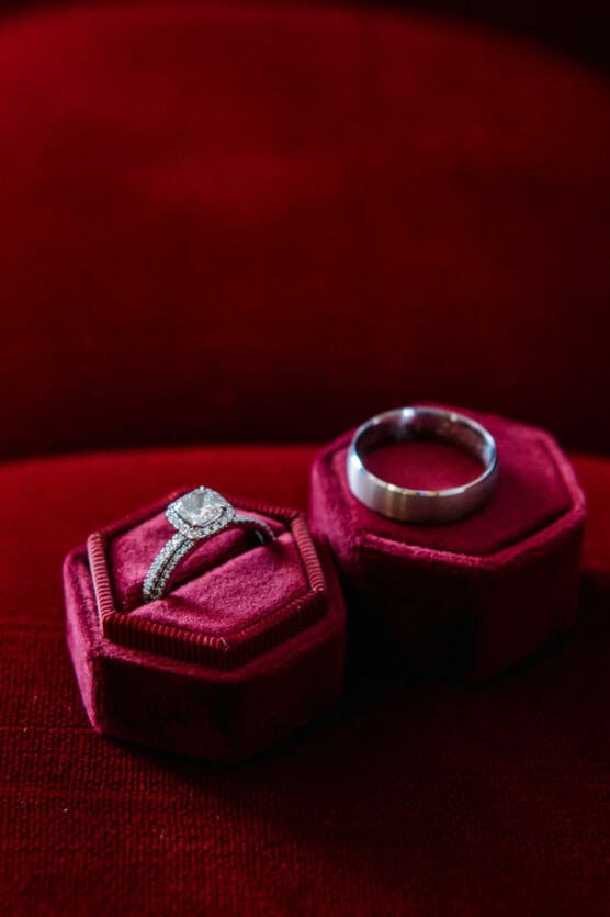 Two rings in red velvet hexagonal boxes on a matching red surface: a diamond engagement ring with a halo setting on the left, and a plain silver wedding band on the right. by Justin Salem Meyer