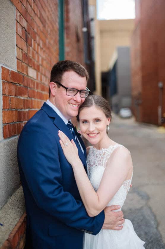A bride in a white lace dress and a groom in a navy blue suit smile and embrace while standing close together in an outdoor urban alleyway with brick walls. by Justin Salem Meyer