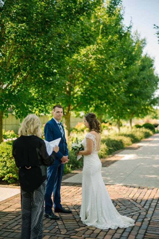 A bride and groom stand holding hands during an outdoor wedding ceremony, facing each other, as an officiant reads to them. They are surrounded by lush green trees and sunlight filters through the leaves. by Justin Salem Meyer