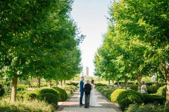 A bride and groom stand facing each other with an officiant between them on a sunlit garden path, surrounded by lush green trees and bushes. A guest stands to the right, taking a photo. by Justin Salem Meyer