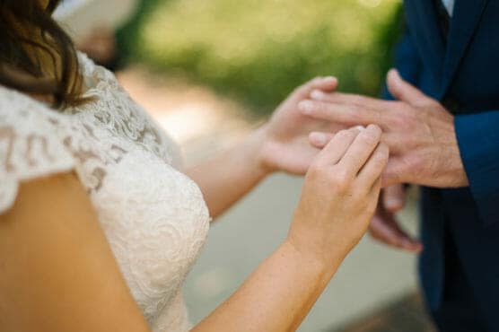 Close-up of a bride in a lace dress placing a ring on the grooms finger during an outdoor wedding ceremony, with greenery blurred in the background. by Justin Salem Meyer