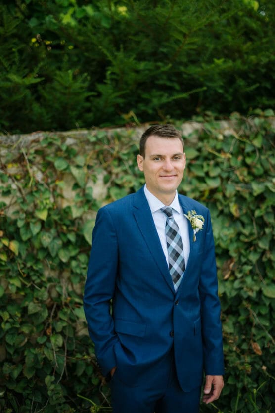 A man in a blue suit with a plaid tie stands in front of a green wall covered in ivy, smiling slightly. He wears a boutonniere on his left lapel. by Justin Salem Meyer