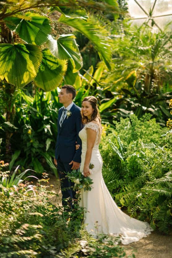 A bride in a white lace gown and groom in a navy suit stand together in a lush garden, surrounded by green tropical plants and sunlight filtering through the leaves. The bride holds a small bouquet and smiles at the camera. by Justin Salem Meyer