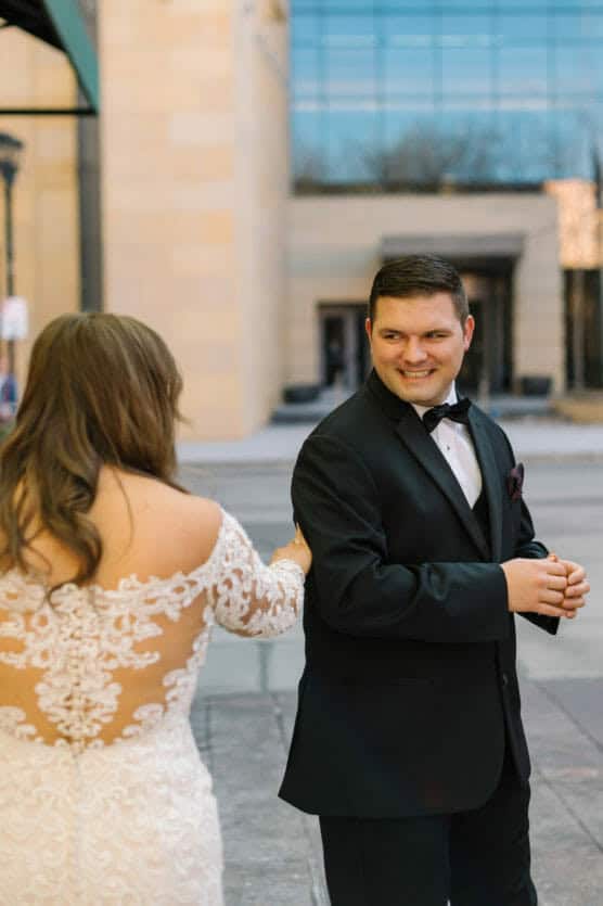 A bride in a lace wedding dress gently holds the arm of a groom in a black tuxedo. The groom smiles warmly at her on a city sidewalk, with tall buildings and reflections in the background. by Justin Salem Meyer