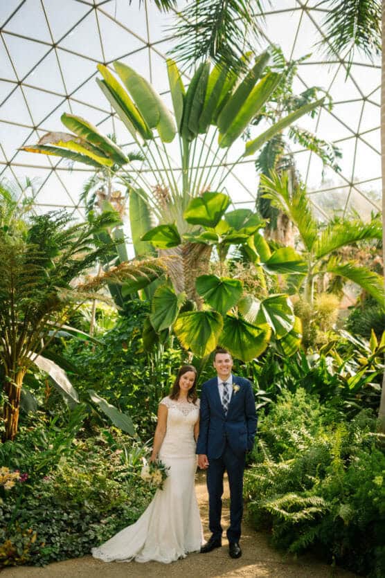A bride in a white dress and a groom in a blue suit stand holding hands, smiling, surrounded by lush tropical plants inside a sunlit greenhouse with a glass dome ceiling. by Justin Salem Meyer