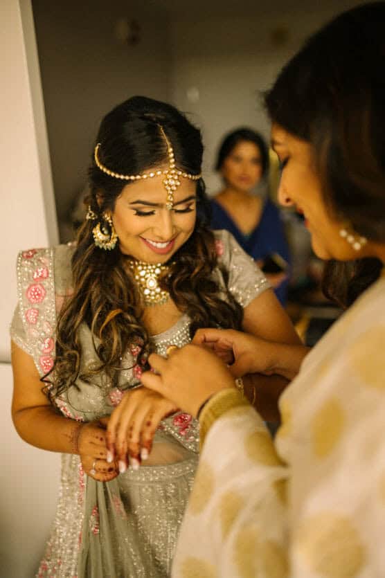 A smiling woman in a silver and pink lehenga with traditional jewelry is getting her hand decorated by another woman in a white and gold saree. Both appear happy in a warmly lit room. by Justin Salem Meyer