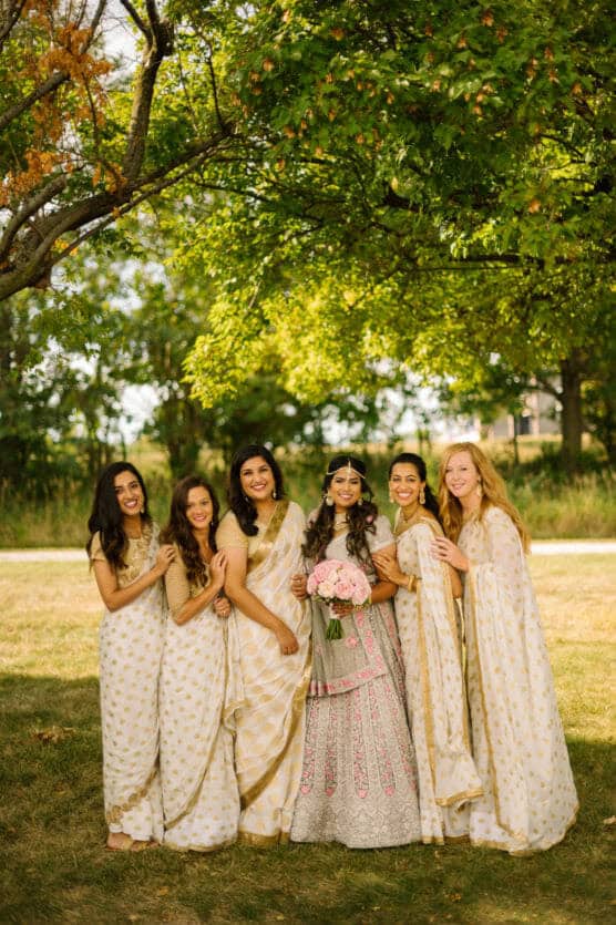 A bride in an ornate lehenga holds a pink bouquet and stands smiling with five bridesmaids in coordinating cream and gold sarees, all posing together outdoors under green trees. by Justin Salem Meyer
