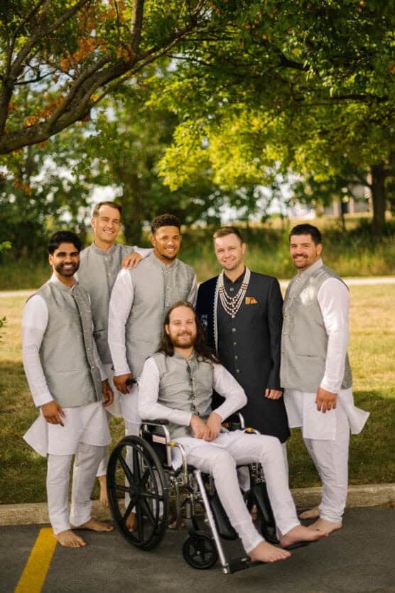 Six men in matching light gray vests and white traditional attire pose together outdoors. One man sits in a wheelchair; another wears a black sherwani. They are all smiling under a tree on a sunny day. by Justin Salem Meyer