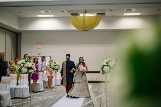 A bride and groom in traditional South Asian attire walk down the aisle together in a decorated indoor wedding venue, surrounded by guests and floral arrangements. by Justin Salem Meyer