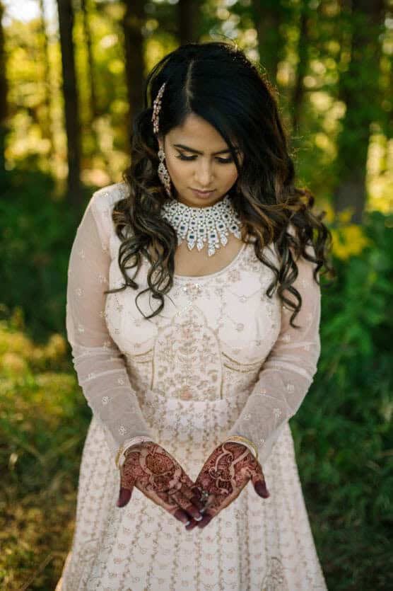 A woman in an embroidered, light-colored dress stands outdoors, looking down at her decorated hands with henna designs. She wears statement jewelry and has long, wavy hair, surrounded by greenery and sunlight filtering through trees. by Justin Salem Meyer