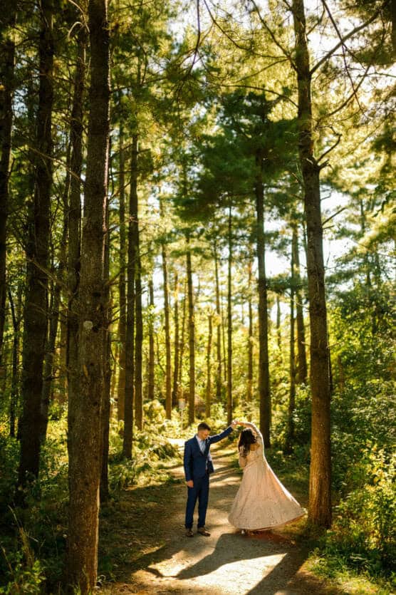 A couple dances on a sunlit forest path; the man wears a blue suit and the woman wears a long, light-colored dress. Tall trees surround them, with sunlight streaming through the branches. by Justin Salem Meyer