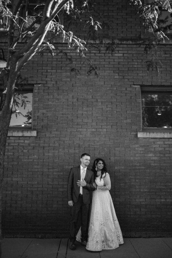 A couple stands close together in front of a brick wall beneath a tree. The man wears a suit and the woman wears a long, patterned dress. They both look toward the camera, framed by two windows in the background. by Justin Salem Meyer