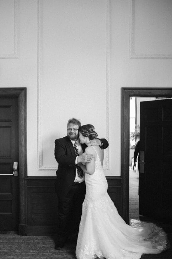 A bride in a lace wedding gown shares a warm embrace with a smiling man in a suit, standing between two doorways in a softly lit, elegant room. The image is in black and white. by Justin Salem Meyer