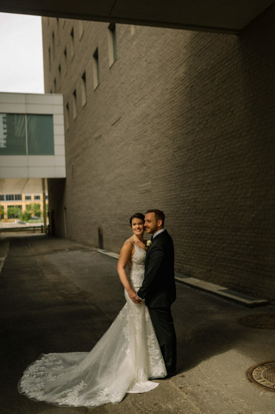 A bride in a white lace gown and a groom in a black suit stand close together, smiling, in a sunlit alley between tall buildings with a skybridge in the background. by Justin Salem Meyer