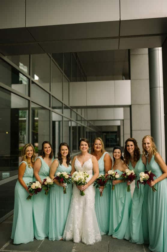 A bride in a white lace gown stands with six bridesmaids in matching light teal dresses, all holding bouquets, smiling in front of a modern building with large windows. by Justin Salem Meyer