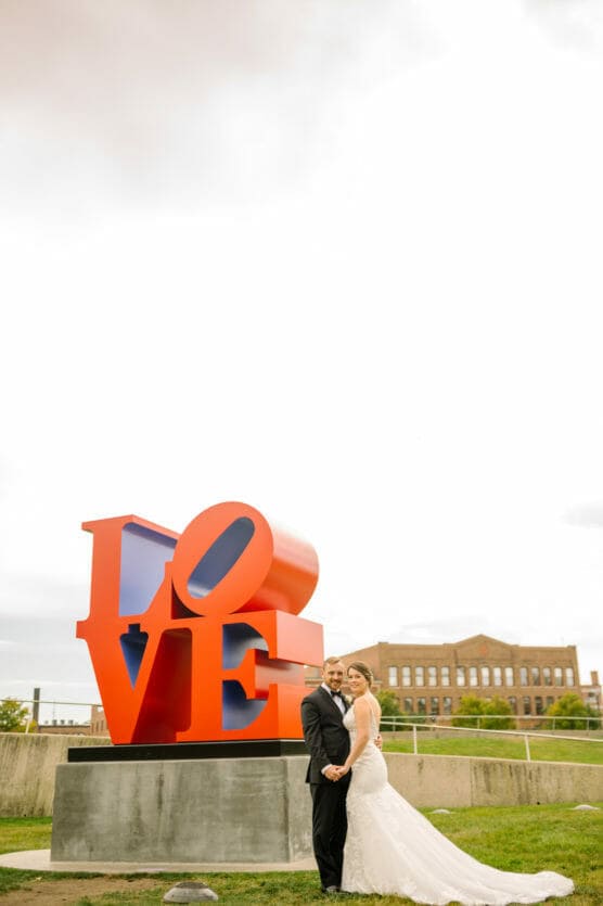A bride and groom stand hand in hand, smiling in front of a large, red and blue LOVE sculpture outdoors, with a historic brick building visible in the background. by Justin Salem Meyer