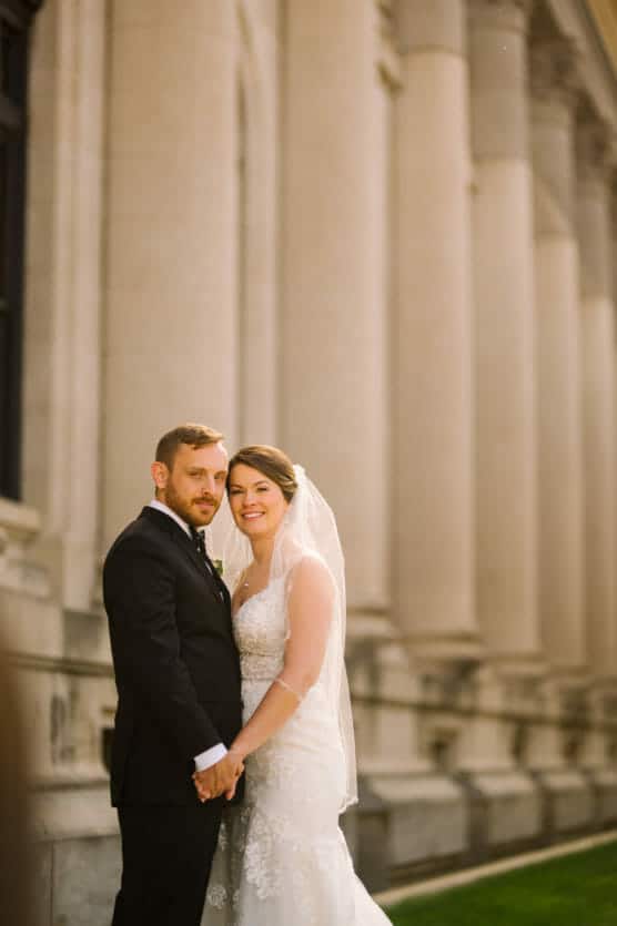 A bride in a white lace gown and veil stands holding hands with a groom in a black suit, both smiling, in front of a grand stone building with tall columns. by Justin Salem Meyer