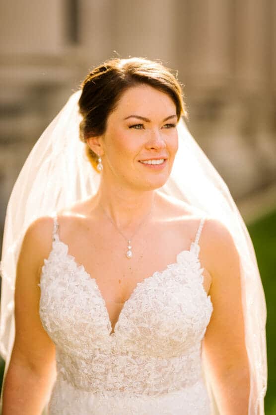 A bride in a lace wedding dress and veil stands outdoors, smiling softly in warm sunlight. She has dark hair styled up and wears pearl earrings and a necklace. The background is softly blurred. by Justin Salem Meyer