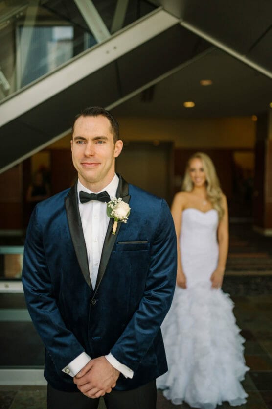 A groom in a blue tuxedo stands in the foreground, smiling, while a bride in a white strapless gown stands behind him, slightly out of focus, in an indoor setting with modern architecture. by Justin Salem Meyer