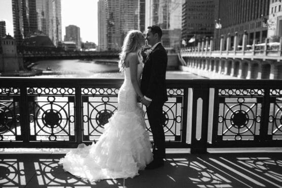 A bride and groom stand face to face on a city bridge, dressed in wedding attire. Tall buildings and a river appear in the background, with sunlight casting shadows through the ornate railing. by Justin Salem Meyer