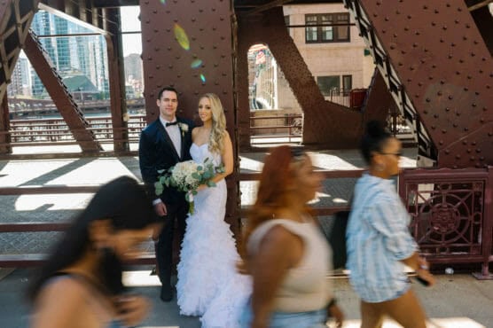 A bride and groom pose for a photo on a city bridge, standing close together and holding a bouquet, while several people walk by in the foreground, slightly blurred from motion. by Justin Salem Meyer