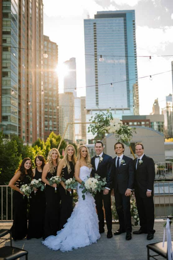 A wedding party poses outdoors on a city rooftop at sunset, with tall buildings in the background. The bride wears a white gown, while bridesmaids wear black dresses and groomsmen wear black suits. Everyone is smiling. by Justin Salem Meyer