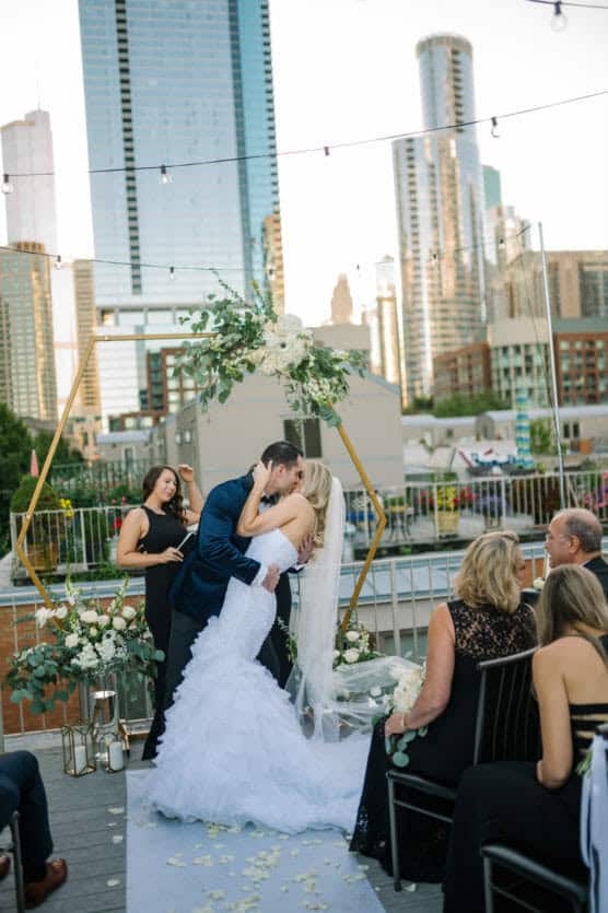 A bride and groom kiss at an outdoor rooftop wedding ceremony, surrounded by guests and city skyscrapers. A geometric arch with flowers decorates the altar, and the officiant stands to the side smiling. by Justin Salem Meyer