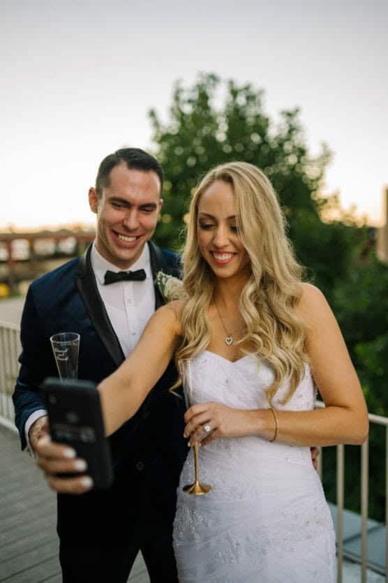 A newlywed couple in formal attire smiles and takes a selfie outdoors. The bride, in a white dress, holds a champagne flute, while the groom, in a dark suit with a bow tie, stands beside her. Trees and a sunset sky are in the background. by Justin Salem Meyer