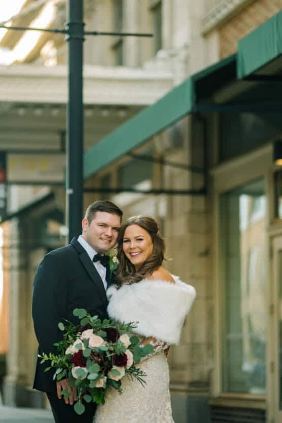 A bride in a white gown and fur shawl stands next to a groom in a black tuxedo. They smile and embrace on a city sidewalk, holding a bouquet with white and dark red flowers. by Justin Salem Meyer