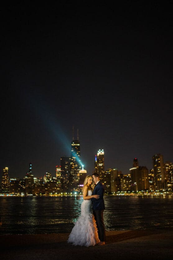 A bride and groom embrace at night in front of a city skyline with illuminated skyscrapers, their reflections shimmering on the water behind them. A bright light projects into the sky from one of the buildings. by Justin Salem Meyer