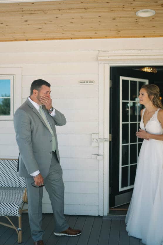 A man in a gray suit appears emotional, covering his mouth with his hand as he looks at a smiling bride in a white dress standing in a doorway. They are outside a white building with wood trim. by Justin Salem Meyer