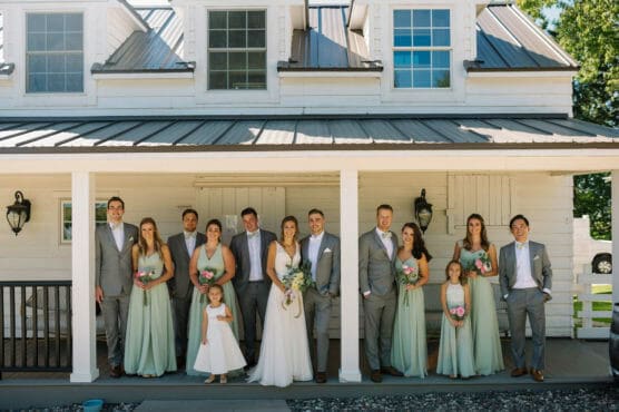 A wedding party poses on a porch in front of a white house. The bride and groom stand in the center, surrounded by bridesmaids in sage green dresses and groomsmen in gray suits. Two flower girls in white dresses hold bouquets. by Justin Salem Meyer