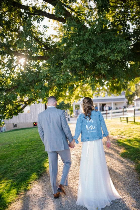 A bride and groom walk hand in hand down a tree-lined path. The bride wears a white dress and a blue denim jacket that says Mrs. Koski. Sunlight shines through the leaves, creating a warm, romantic scene. by Justin Salem Meyer