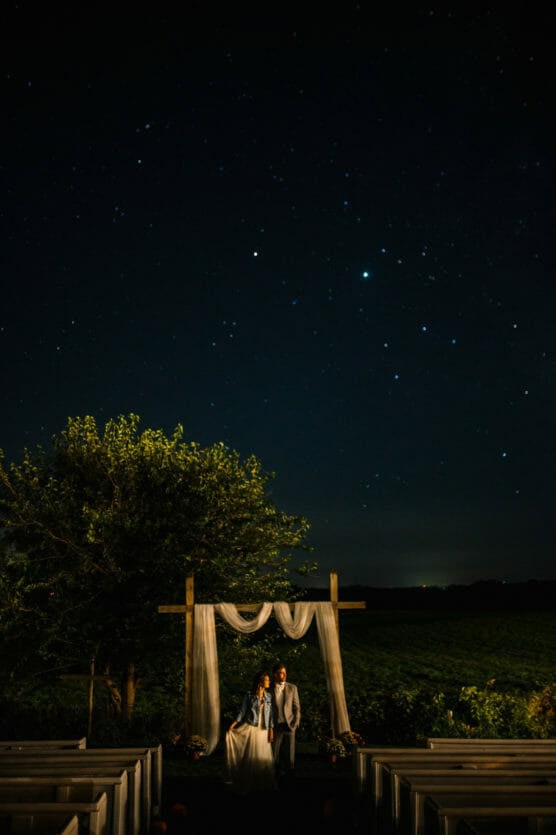 A bride and groom stand under a wooden wedding arch draped with white fabric, outdoors at night, beneath a star-filled sky and surrounded by empty benches and greenery. by Justin Salem Meyer