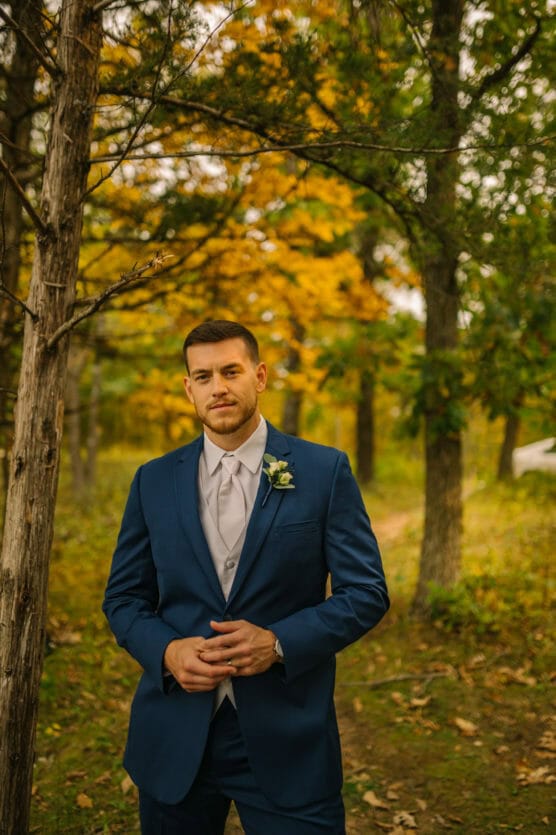 A man in a blue suit and light gray tie stands outdoors in a forest with autumn foliage, looking at the camera with a slight smile. The background features trees with green and yellow leaves. by Justin Salem Meyer