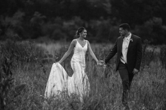 A bride in a white gown and a groom in a suit walk hand-in-hand through a grassy field, smiling at each other, with trees blurred in the background. by Justin Salem Meyer